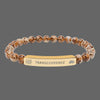 A beaded stretch bracelet featuring brown and beige marbled beads with a central gold-toned metal ID plate engraved with the word 'TRANSCENDENCE' and small spiritual symbols.