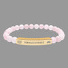 A pink beaded bracelet with a gold-toned rectangular ID plate engraved with the word 'TRANSCENDENCE' and small galaxy spiral and lotus flower symbols on either side.
