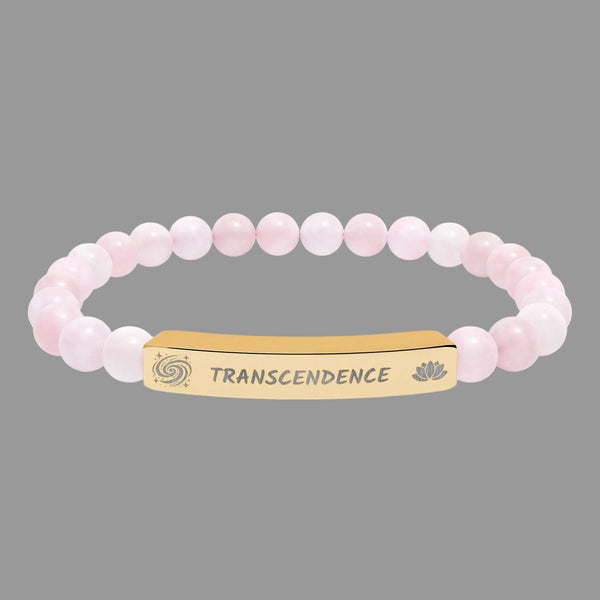 A pink beaded bracelet with a gold-toned rectangular ID plate engraved with the word 'TRANSCENDENCE' and small galaxy spiral and lotus flower symbols on either side.