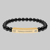 A beaded stretch bracelet featuring glossy black round beads and a central gold-toned metal ID plate engraved with the word 'TRANSCENDENCE' and small spiritual symbols on either side.