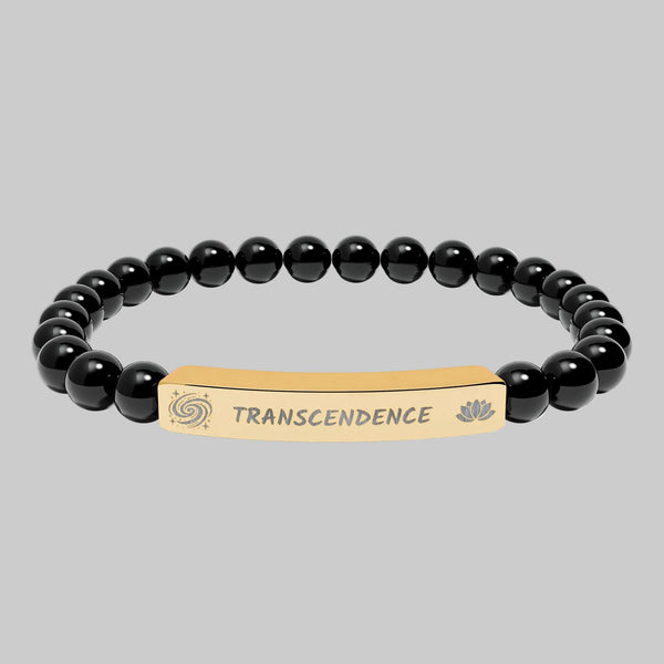 A beaded stretch bracelet featuring glossy black round beads and a central gold-toned metal ID plate engraved with the word 'TRANSCENDENCE' and small spiritual symbols on either side.