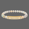 A beaded stretch bracelet featuring a central gold-toned bar engraved with the  spiritual word 'TRANSCENDENCE' and small lotus and spiral motifs. The beads are a mix of soft pastel tones including cream, peach, pale green, and light gray.