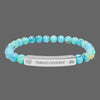 A beaded stretch bracelet featuring light blue and turquoise marbled beads with black veining, connected by a polished silver metal ID plate engraved with the word 'TRANSCENDENCE' and spiritual symbols of galaxy and lotus flower.