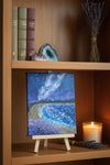 Painting of a night sky with Milky Way galaxy reflected on a beach scene with waves and purple flowers, artist's signature visible.