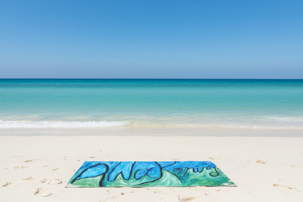 A rectangular beach towel featuring an abstract, swirling art design in shades of blue and green with 'Awakening' in black ink script, displayed flat on a sansy beach.